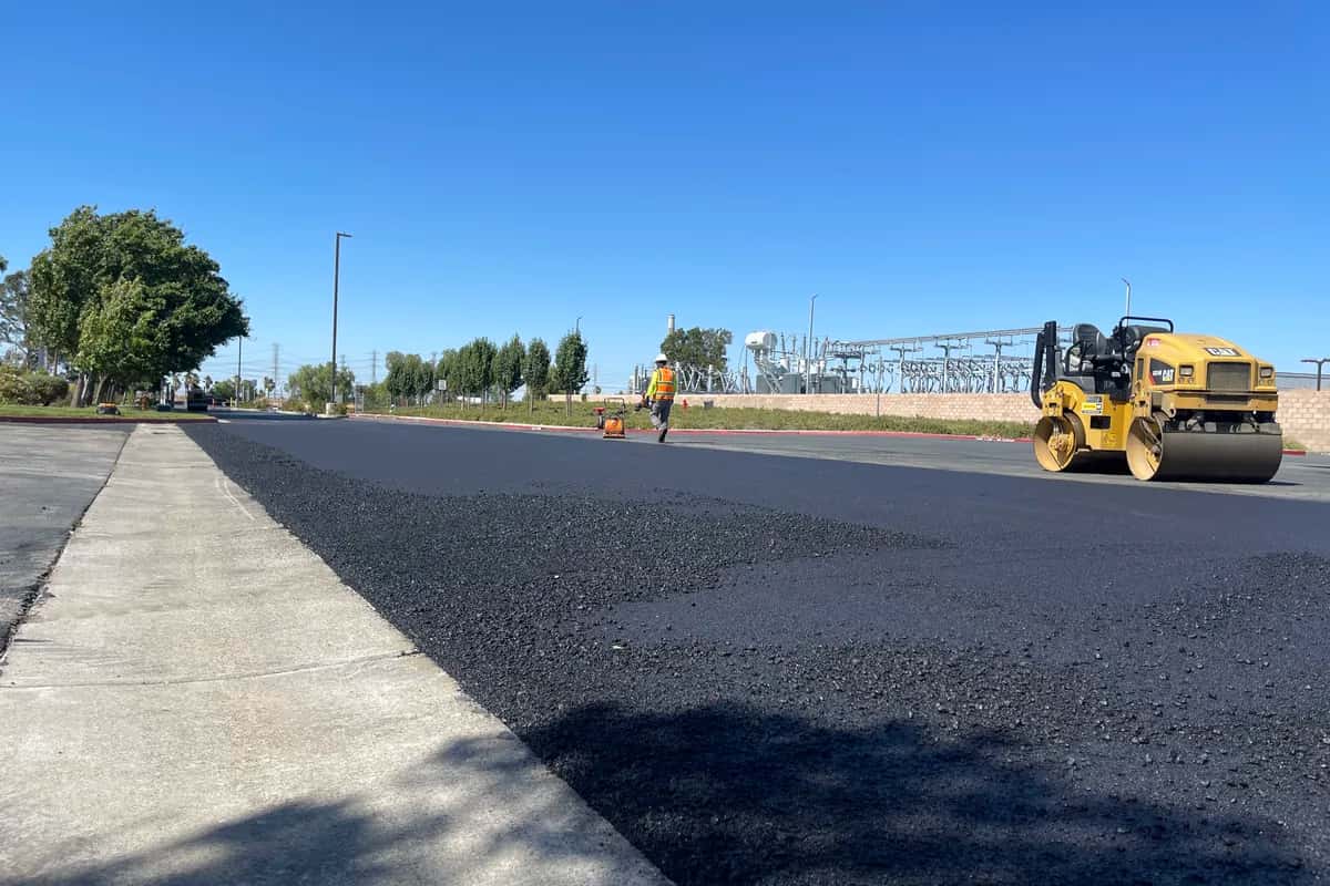 Roller compacting a freshly placed asphalt mat next to a concrete walkway on a commercial paving project.